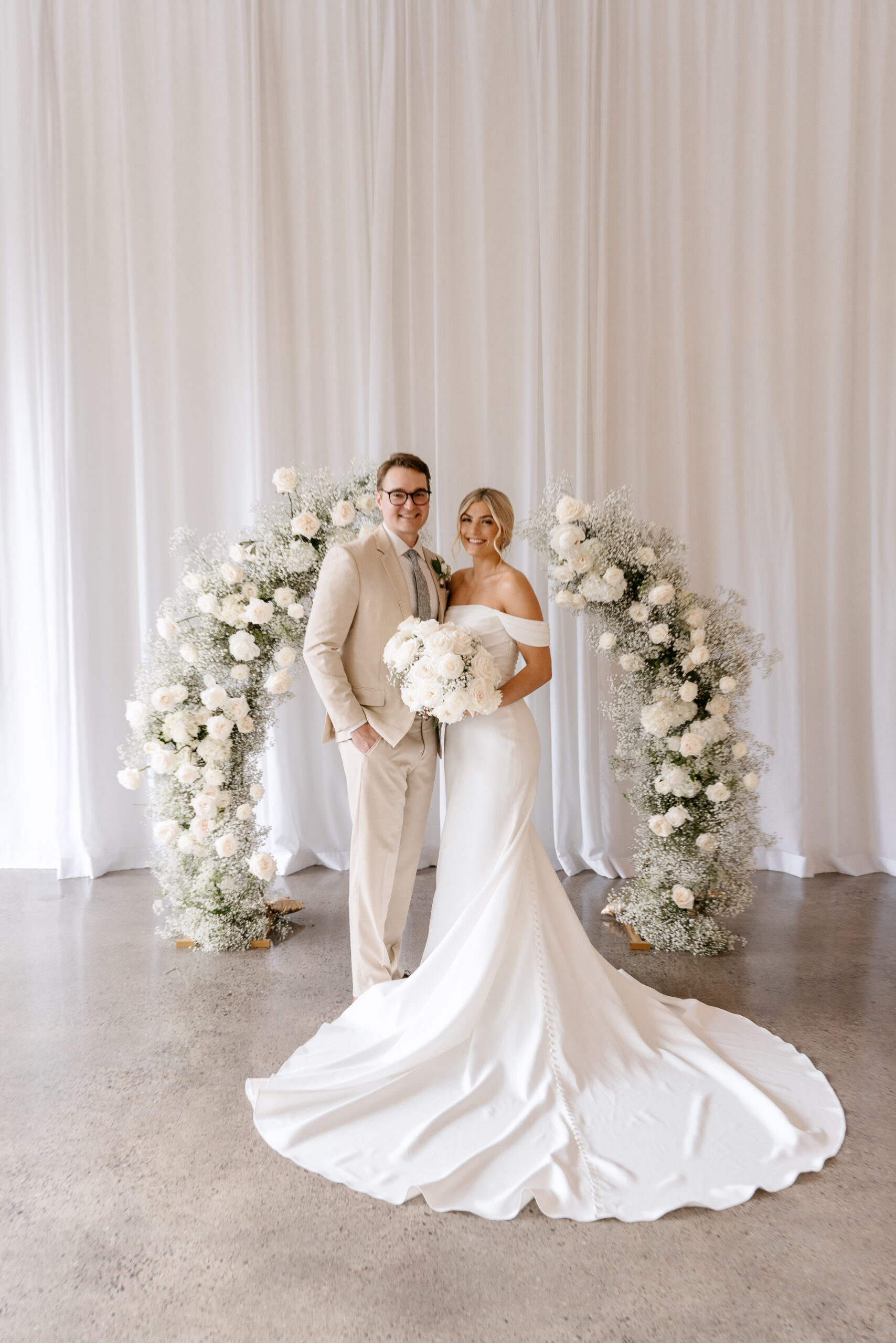 bride and groom in front of white floral arbor
